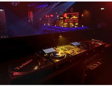 DJ-Setup in einem dunklen Club mit Mischpult, Plattenspielern und beleuchteten Kontrollen im Vordergrund. Im Hintergrund ist eine Bar mit roten und blauen Lichtern erkennbar.