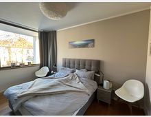 Ein modernes Schlafzimmer mit einem grauen Polsterbett, flankiert von Nachttischen mit Lampen. Ein großes Fenster mit grauen Vorhängen bietet Blick auf eine helle Außenfassade, und an der Wand hängt ein Landschaftsfoto.