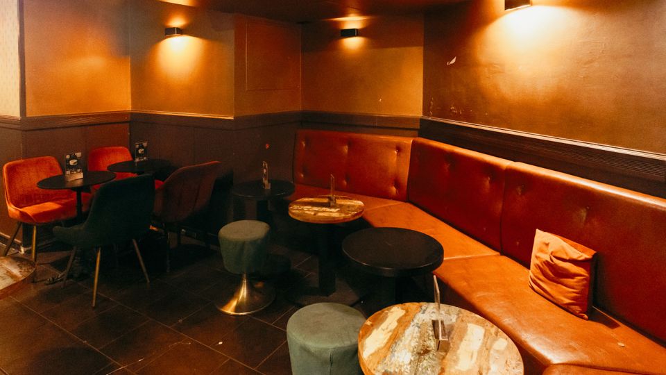 Gemütlicher Bar- oder Lounge-Innenraum mit langen, gepolsterten Sitzbänken aus rotem Kunstleder entlang der Wand, mehreren kleinen runden Tischen und Hockern. Warme Wandlampen und dunkel geflieste Böden prägen die gedämpfte Beleuchtung und das intime Ambiente.