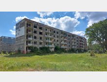 Ein verlassenes, mehrstöckiges Wohngebäude mit beschädigten Fassaden und Balkonen, teilweise mit Graffiti bedeckt. Das Gebäude steht inmitten einer verwilderten Grünfläche, umgeben von Bäumen und vereinzelten Büschen unter einem wolkigen Himmel.