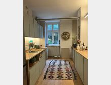 Moderne Küche mit hellgrauen Schränken und Arbeitsplatten aus Holz. Der Boden ist aus Holz, darüber liegt ein bunt gemusterter Teppich, während ein großes Fenster mit Blick ins Grüne den Raum erhellt; die Wände bestehen aus weiß gestrichenen Ziegelsteinen.