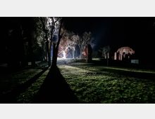 Nachtszene mit Ruinen einer roten Backsteinarchitektur auf einer Wiese, umgeben von Bäumen, die von hellem Licht im Hintergrund beleuchtet werden. Lange Schatten der Bäume fallen über das grasbewachsene Gelände, und die Umgebung wirkt dunkel bis auf die erleuchtete Ruine.