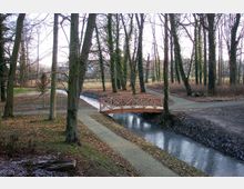 Ein Park mit einem kleinen Bach, der von einer hölzernen Fußgängerbrücke überspannt wird, umgeben von hohen, teils mit Efeu bewachsenen Bäumen und Wegen. Der Boden ist mit Laub bedeckt, und die Vegetation wirkt winterlich kahl.