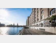 Uferpromenade mit Kopfsteinpflaster, flankiert von einem historischen Backsteingebäude mit großen Fenstern. Rechts stehen überdachte Sitzbereiche mit weißen Stoffpavillons, links fließt ein Fluss mit der Oberbaumbrücke im Hintergrund, beleuchtet von der Sonne.