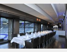 Innenraum einer modernen Lounge mit Blick auf ein Stadion. Der Raum ist mit einer langen, festlich gedeckten Tafel, schwarzen Lederstühlen, großer Fensterfront und einer Bar im Hintergrund gestaltet.