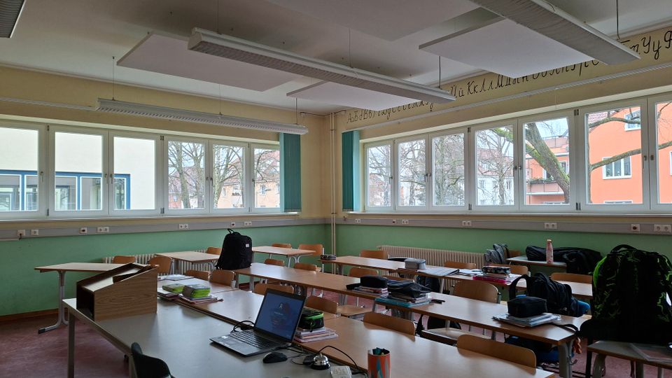 Klassenzimmer mit mehreren Reihen aus Holztischen und Stühlen sowie einem Lehrerpult im Vordergrund. Große Fensterfronten mit Blick auf Bäume und umliegende Gebäude, grün- und beigefarbene Wände und abgehängte Deckenpaneele mit Leuchtstofflampen prägen den Raum.