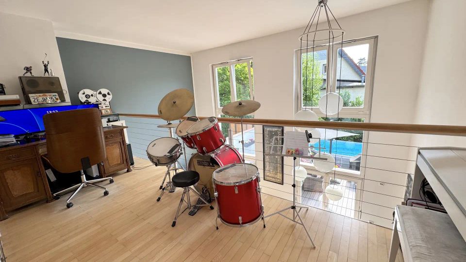 Offener Galerieraum in einem Wohnhaus mit hellem Holzboden, großem Fenster und Geländer mit Holzhandlauf über einer Treppe. Im Vordergrund steht ein rotes Schlagzeug, daneben hängen mehrere runde Lampenschirme an einer Pendelleuchte.