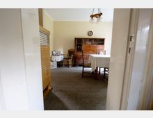 Blick durch eine Tür in ein schlichtes Esszimmer mit grauem Teppichboden, einem Esstisch mit weißer Tischdecke und mehreren Holzstühlen. Im Hintergrund stehen eine dunkle Holzvitrine und ein kleiner Beistelltisch mit Dekoration; an der Decke hängt eine zweiflammige Leuchte.