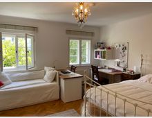 Helles Schlafzimmer mit Holzparkettboden, einem weißen Tagesbett links, einem Schreibtisch mit Regal sowie Pinnwand in der Mitte und einem weißen Metallbett rechts. Große Fenster mit Blick ins Grüne lassen viel Tageslicht herein, und eine klassische Deckenlampe hängt zentral im Raum.