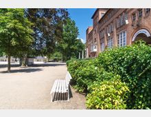 Backsteinfassade eines Gebäudes mit großen Fenstern und einem Bogeneingang, daneben eine Hecke mit Blumen und eine weiße Parkbank im Vordergrund. Im Hintergrund sind Bäume, ein sonniger Platz und kleine Gebäude erkennbar.