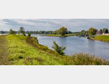Landschaft mit einem Fluss, der sich durch eine grüne, von Gras und vereinzelten Bäumen bewachsene Uferlandschaft schlängelt; im Hintergrund ein kleines Dorf mit roten Ziegeldächern und einem Anleger. Der Himmel ist teils bewölkt, und die Umgebung wirkt ruhig.