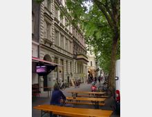 Straßenszene mit Altbaufassaden und einem Baum gesäumten Bürgersteig, auf dem Fahrräder abgestellt und Holzbänke sowie Tische aufgestellt sind. Im Vordergrund sitzt eine Person, während im Hintergrund Passanten und Lokalschilder zu sehen sind.