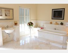 Helles Badezimmer mit cremefarbenen Marmorelementen, einer freistehenden Badewanne und einem kleinen Waschbecken. Ein großes Fenster mit Fensterläden lässt Tageslicht herein, daneben steht ein dekorativer Blumenstrauß auf einer Säule.