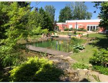 Ein Garten mit einem naturbelassenen Teich, umgeben von üppiger Vegetation und Pflanzen. Im Hintergrund steht ein rosafarbenes, einstöckiges Haus mit großen Fenstern und einer Terrasse mit Sitzgelegenheiten und Sonnenschirm.