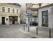 Straßenszene in einem historischen Viertel mit Kopfsteinpflaster. Links ein gelbes Gebäude mit der Aufschrift "Kietzer Bäckerei" und großen Schaufenstern, rechts ein graues Geschäft mit einem Schild "Uhren & Schmuck", im Hintergrund weitere Altbauten.