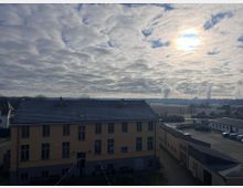 Ein zweistöckiges gelbes Gebäude mit grauem Dach in ländlicher Umgebung, umgeben von weiteren Gebäuden und einer gepflasterten Fläche. Im Hintergrund eine weite Landschaft mit Feldern, Bäumen und aufsteigendem Dampf unter einer bewölkten Himmelsszenerie mit durchscheinender Sonne.