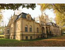 Historisches Herrenhaus aus gelbem Stein mit neogotischen Elementen, darunter spitz zulaufende Türmchen, Rundbogenfenster und ein großer Eingangsbereich mit Säulen; umgeben von herbstlichem Laub und Bäumen in einem parkähnlichen Gelände.