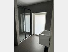 Modernes Badezimmer mit dunklen Fliesenboden und grauen Wänden, einer bodenebenen Dusche mit Glasschiebetür sowie einem kleinen weißen Waschbecken gegenüber einer matten Glastür.