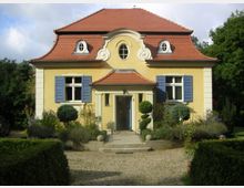 Gelbes Einfamilienhaus mit rotem Walmdach und blauen Fensterläden, symmetrisch gestaltet mit einem zentralen Eingang, flankiert von Säulen und kleinen Zierbäumen. Im Vordergrund befindet sich ein Kiesweg, umgeben von gepflegter Hecken- und Blumenbepflanzung.