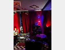 Ein kleiner, gemütlicher Raum mit roten Vorhängen, schwarz-weiß kariertem Boden und gedämpfter Beleuchtung. Im Raum befinden sich runde Holztische mit Stühlen, ein Regal mit Büchern und Dekoobjekten sowie eine Lampe mit rotem Lampenschirm.