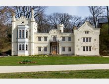 Schlossähnliches Gebäude im neogotischen Stil mit cremefarbener Fassade, Zinnen, Spitzbogenfenstern und einem Erker auf der linken Seite. Das Gebäude steht vor einer Rasenfläche mit Blumenbeeten, umgeben von kahlen Bäumen.