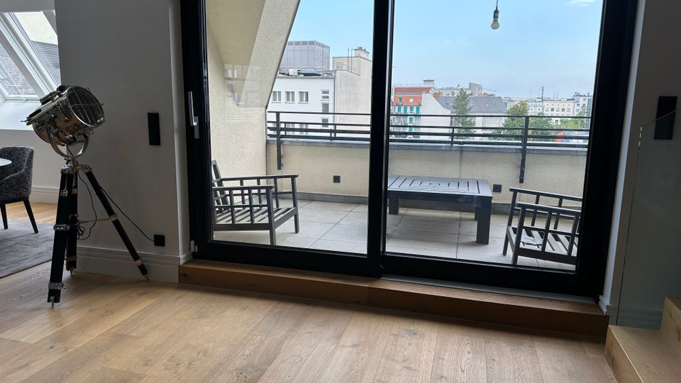 Blick aus einem modernen Wohnraum mit Holzfußboden durch eine bodentiefe Glas-Schiebetür auf eine Dachterrasse mit Fliesen, Sitzmöbeln und Geländer. Im Hintergrund ist eine städtische Gebäudekulisse unter blauem Himmel zu sehen.
