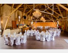 Ein großer Veranstaltungsraum mit Backsteinwänden, Holzgebälk und dekorativen Kronleuchtern. Die Tische sind mit weißen Hussen, Geschirr und Servietten eingedeckt, und Lichterketten schmücken die Balken.