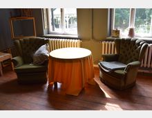 Zwei grüne, gepolsterte Sessel stehen gegenüber einem runden Tisch mit gelber Tischdecke in einem Raum mit Holzdielen. Im Hintergrund befinden sich ein Fenster mit Blick nach draußen, eine Heizung und mehrere Dekorationsgegenstände.