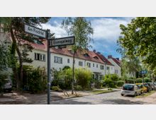 Straßenszene in einer Wohngegend mit weiß verputzten Reihenhäusern und roten Dächern, umgeben von Bäumen und Gärten. Im Vordergrund stehen Straßenschilder mit den Namen „Am Fischtal“ und „Eisvogelweg“ sowie parkende Autos entlang einer gepflasterten Straße.