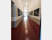 Ein langer, heller Flur mit roten Fußböden, beige-weißen Wänden und großen Fenstern am Ende. An den Wänden hängen Luftaufnahmen, und runde Deckenleuchten hängen gleichmäßig entlang der Decke.