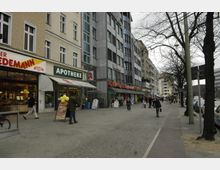Straßenszene mit mehreren Geschäften in einem urbanen Umfeld, darunter eine Bäckerei und eine Apotheke, erkennbar durch Ladenschilder. Links sind ältere Gebäude mit hellen Fassaden und rechts moderne, gläserne Gebäude; Passanten und geparkte Autos säumen den Gehweg.
