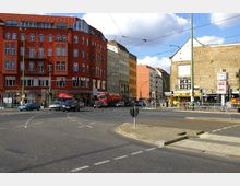 Straßenkreuzung in einer Stadt mit mehrstöckigen Wohn- und Geschäftshäusern, darunter ein rotes Gebäude mit großen Fenstern und Balkonen. Im Vordergrund Straßenmarkierungen, Autos, Straßenbahnschienen und Fußgänger; im Hintergrund ein Mauerwerk mit Graffiti und diverse Schilder.
