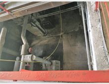 Blick in einen technischen Schacht mit Rohrleitungen und einem roten Absperrventil. Der Bereich ist umgeben von Betonwänden und teilweise durch eine rote Metallabdeckung eingerahmt.