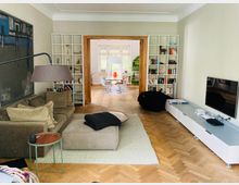 Wohnzimmer mit einem großen beigen Sofa, einer Stehleuchte und einem Teppich, daneben Regale mit Büchern. Im Hintergrund ein angrenzender Essbereich mit großen Fenstern, hellem Holzfußboden und einer modernen Esstischgruppe.