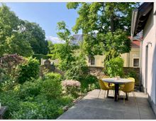 Hof mit Garten und Terrasse, umgeben von grüner Vegetation und blühenden Sträuchern. Im Vordergrund steht ein runder Tisch mit vier gelben Stühlen auf einer gepflasterten Fläche, im Hintergrund sieht man eine gelbe Hauswand mit Solarpanelen auf dem Dach.