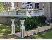 Eine Terrasse mit weißem Geländer und dekorativen Blumenkästen, die auf Natursteinstützen ruht. Im Vordergrund ein gepflasterter Gartenbereich mit weißen Pflanzgefäßen und grünen Sträuchern, umgeben von Rasen und Blumenbeeten.