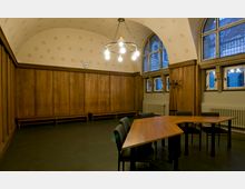 Ein historisch eingerichteter Besprechungsraum mit gewölbter Decke, Holztäfelungen an den Wänden und hohen Fenstern mit Bleiverglasung. Im Raum stehen trapezförmige Tische mit Stühlen und ein Kronleuchter beleuchtet die Szene.