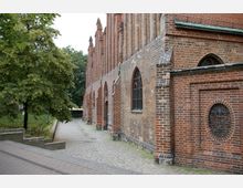 Seitenansicht einer Backsteinkirche mit gotischen Spitzbogenfenstern und dekorativen Mauerdetails. Ein gepflasterter Weg führt entlang der Kirchenwand, flankiert von einem Baum und Grünflächen.