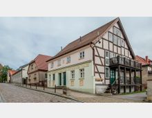 Fachwerkhaus mit zweigeschossigem Balkon und rotem Satteldach, Teil einer gepflasterten Straßenszene mit weiteren historischen Gebäuden im Hintergrund. Auf der Fassade des Hauses steht "Kleines Wartenbergheim zum Historischen Hause".