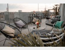 Moderne Dachterrasse mit Sitzgelegenheiten aus schwarzen Metallstühlen und Sofas mit farbigen Kissen. Im Vordergrund ein kleiner runder Tisch mit Blumenarrangement, im Hintergrund eine geländerartige Abgrenzung und Hochhäuser in der Ferne.