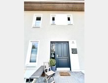 Hauseingang einer modernen Wohnhausfassade mit grauer Tür, Hausnummer 40 und schwarzem Briefkasten. Vor der Tür stehen ein Holzstuhl, eine Holztischbank und eine Pflanze in einem Topf auf gepflastertem Boden.