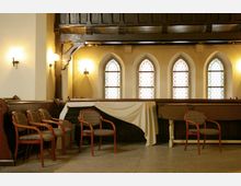 Innenraum einer historischen Kirche mit einer Holzgalerie und vier gotischen Fenstern mit buntem Glas. Im Vordergrund stehen mehrere gepolsterte Holzstühle und ein mit weißem Tuch bedeckter Tisch.