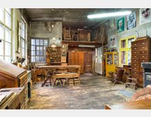 Werkstatt mit rustikalem Holzinterieur, ausgestattet mit Werkbänken, Regalen voller Werkzeuge sowie Schränken und Schubladen. Der Raum verfügt über große Fenster, eine abgenutzte Holzbodendiele und an den Wänden hängen mehrere Porträtbilder.