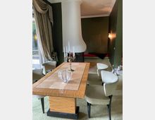 Esszimmer mit einem rechteckigen Holztisch, umgeben von gepolsterten Stühlen. Im Hintergrund ein Kamin mit geschwungener weißer Verkleidung und eine dunkelgrüne Wand, seitlich ein großes Fenster mit schweren Vorhängen.