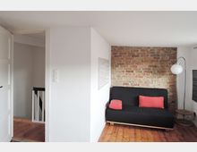 Wohnraum mit einer teilweise freiliegenden Ziegelwand, einem schwarzen Sofa mit roten Kissen, einer hölzernen Bodenfläche und einer Stehleuchte mit weißem Lampenschirm. Links führt eine weiße Tür mit Blick auf eine Treppe in einen angrenzenden Bereich.