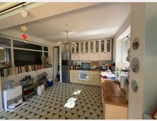 Helle Wohnküche mit pastellgelben Küchenmöbeln, einer Holzarbeitsplatte und Glasoberschränken. Auf der linken Seite stehen Bücherregale, ein Kühlschrank und ein Fenster, das Tageslicht hereinlässt; der Boden ist mit geometrischem Fliesenmuster gestaltet.