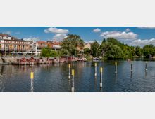 Uferbereich eines Flusses mit mehreren roten Hausbooten an einem Steg, umliegenden Gebäuden mit roten Ziegeldächern und üppigen Bäumen im Hintergrund. Im Wasser stehen gelb markierte Pfosten, und ein kleines Boot ist am rechten Bildrand zu sehen.
