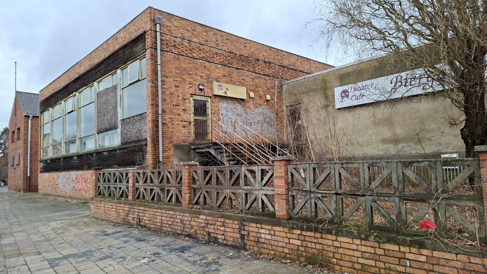 Backsteinbau mit großen Fensterfronten und einem seitlichen Treppenaufgang zu einer vergitterten Tür, davor ein niedriger Ziegel- und Betonzaun entlang eines Gehwegs. Rechts schließt eine verputzte Wand mit einem Schild „Theater Café“ an, daneben kahle Bäume und verwildertes Gelände.