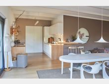 Moderner Wohn- und Essbereich mit offener Küche; weiße Möbel und eine Küchenzeile aus Holz mit Hockern, dekorative Hängeleuchten, ein runder Esstisch mit Stühlen sowie ein graues Sofa auf der rechten Seite. Große Fensterfront links und Holzfußboden.