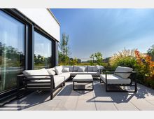 Moderne Terrasse mit hellgrauem Outdoor-Sofa-Set und Glasfront eines Gebäudes im Hintergrund. Umgeben von Pflanzen, darunter Ziergräser und Sträucher, mit Blick auf eine grüne Landschaft unter blauem Himmel.